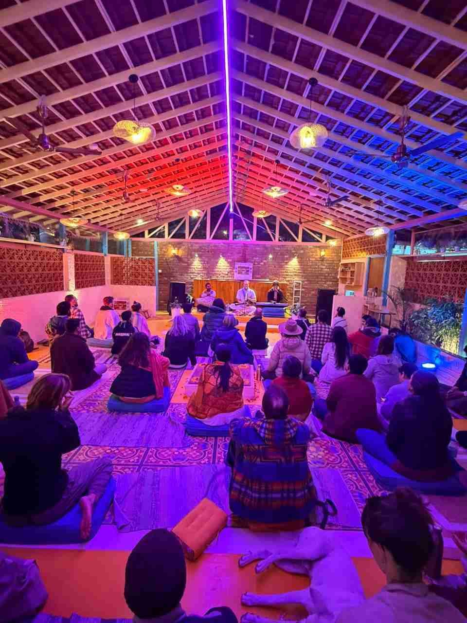 A kirtan gathering in a candlelit hall, people seated on cushions facing the musicians